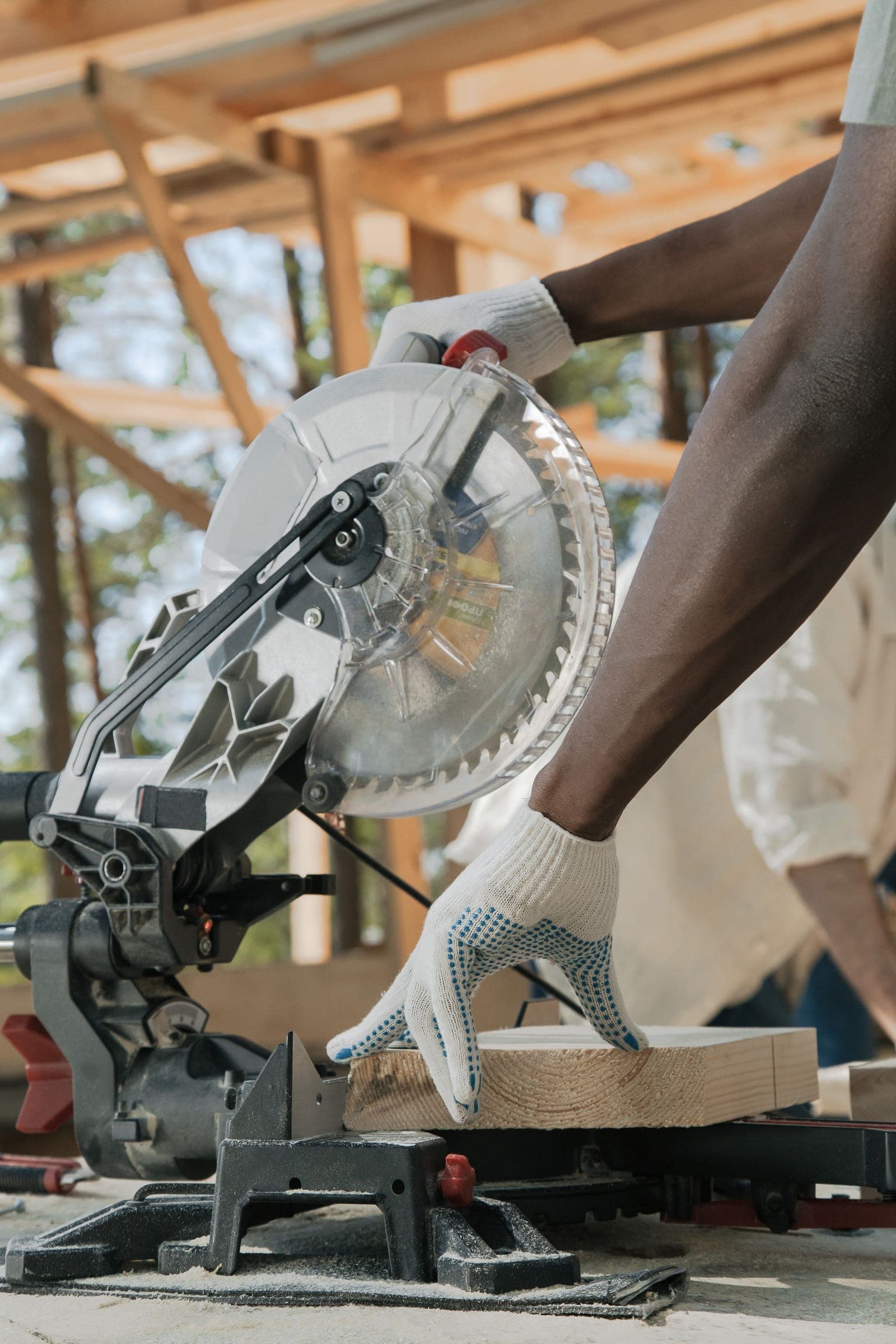 Builder's gloved hands cutting lumber on a miter saw beneath fresh timber framing